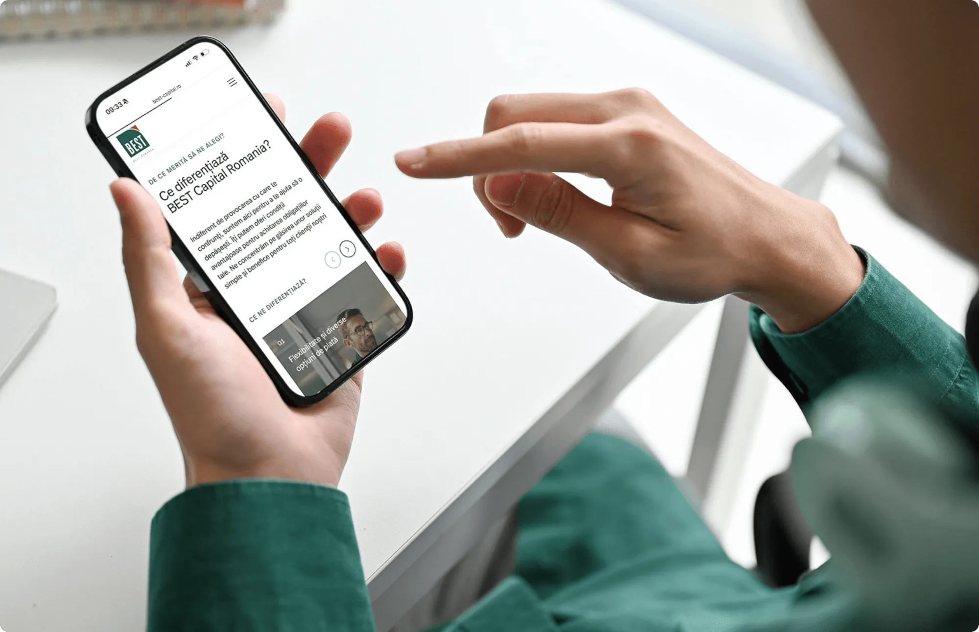 Dłonie trzymające telefon ze stroną best-capital.ro, obok druga dłoń przewija ekran.