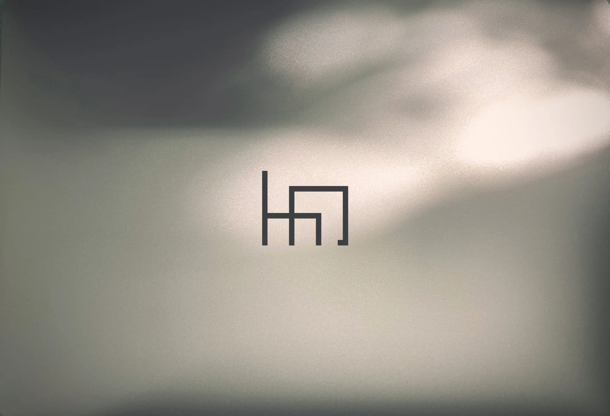 Sygnet Hana Design na jasnoszarej ścianie w miękkim świetle.