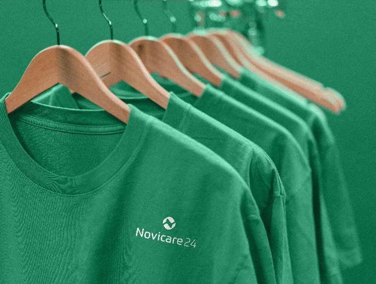 Zielone koszulki z logo Novicare24 na drewnianych wieszakach