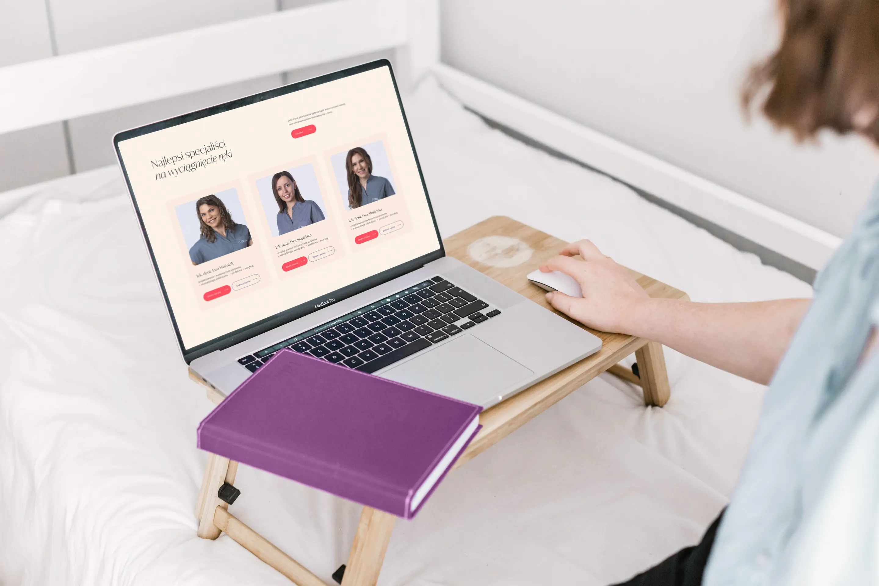 Laptop na stoliku w łóżku, na ekranie lista specjalistów kliniki z przyciskami rezerwacji.