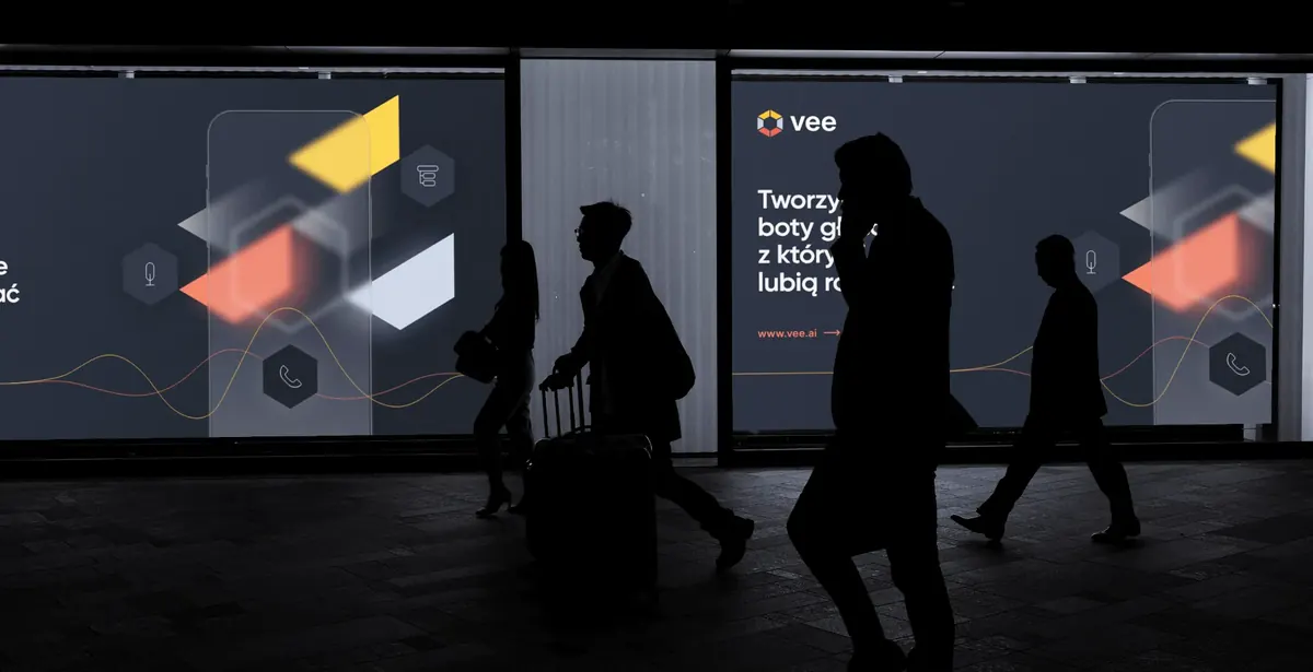 Billboard Vee w przejściu podziemnym z hasłem „Tworzymy boty głosowe, z którymi ludzie lubią rozmawiać”, przed nim sylwetki przechodniów.